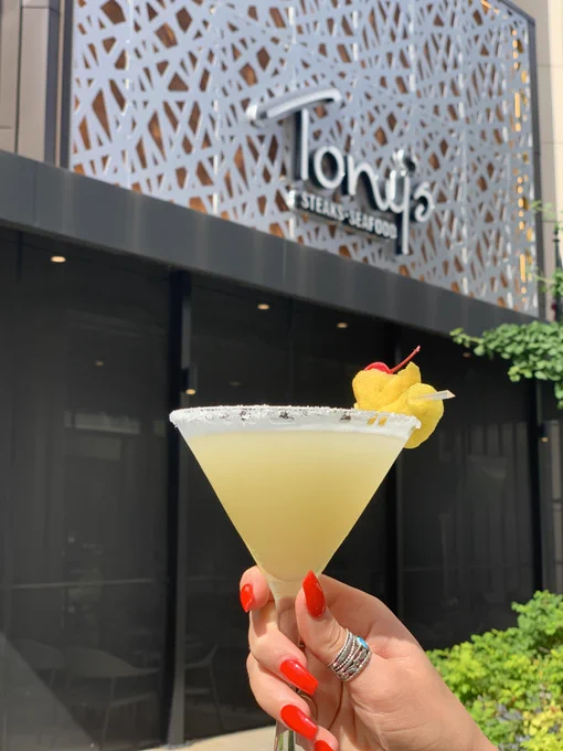 Close up of a woman’s hand with long red nails holding chilled martini glassed garnished with fruit in front of Tony’s of Indianapolis’s exterior sign.