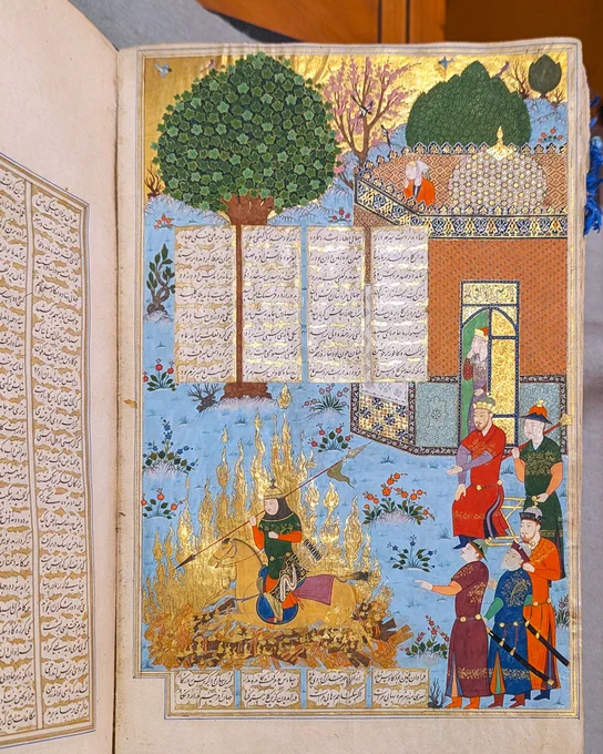 An illuminated manuscript page depicting a historical scene, featuring a person surrounded by golden flames. Several individuals, dressed in vibrant traditional attire, stand nearby gesturing towards the individual. The backdrop includes a lush tree and an ornate building with intricate patterns. The page is adorned with Persian script.