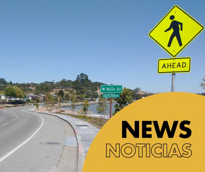 A curved road with a pedestrian crossing sign reading "AHEAD." A street sign indicates "Main Dr." Trees line the road, and hills are visible in the background. A yellow shape in the foreground displays the words "NEWS NOTICIAS.