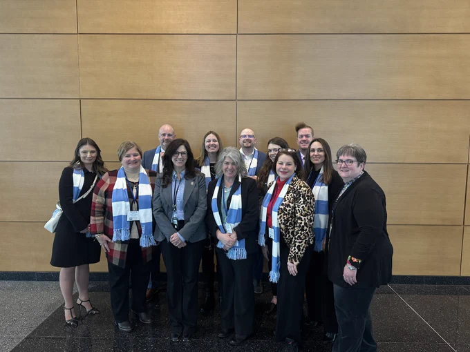 A group of eleven people standing together in a well-lit room with a wood-paneled wall. Some are wearing light blue and white scarves. Everyone is dressed in business attire, smiling at the camera.