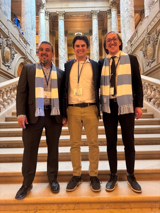 Three people are standing on a grand staircase with ornate pillars in the background, each wearing a light blue and white striped scarf. They are smiling and dressed in semi-formal attire with visible name tags. The setting appears to be a significant historical or governmental building.