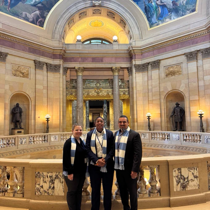 A group of three people stands inside an ornate, classical-style building, possibly a capitol, with tall columns, statues, and a detailed painted dome. They wear blue and white scarves, posing for a photo in the grand interior.
