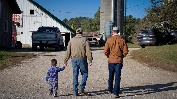 A photo of Rob Sand walking up a gravel drive way towards a farm with a farmer & his son. 
