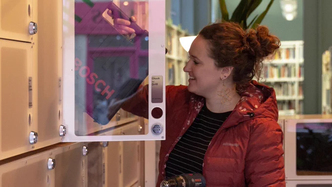 Woman in red jacket retrieves a Bosch drill from a locker in a library setting.