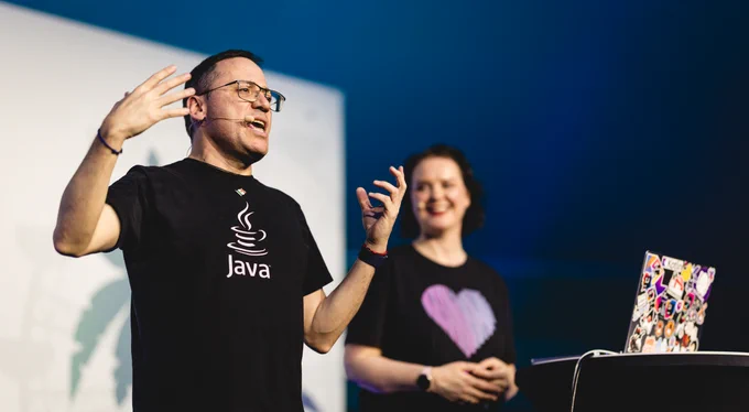 Andres Almiray speaking at JavaLand. He is wearing a black Java t-shirt. Marit van Dijk is in the background looking at him.