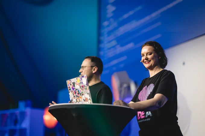 Marit van Dijk standing behind the lectern that holds her laptop. She is smiling at the camera. Andres Almiray is in the background.
