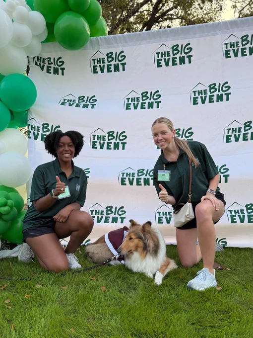 Two people kneel on grass next to Texas A&M mascot, Reveille. They are in front of a backdrop with "The Big Event" logos. There are green and white balloons on the side. Both individuals are smiling at the camera.