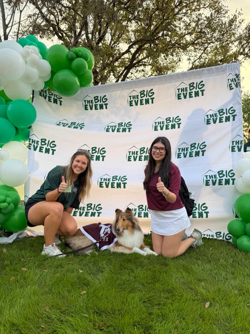 Two people are kneeling on grass with Texas A&M mascot, Reveille, wearing a maroon blanket in front of a backdrop that reads "The Big Event" repeatedly. The backdrop is decorated with green and white balloons. The individuals are posing with one hand showing a thumbs-up gesture. Trees are visible in the background.