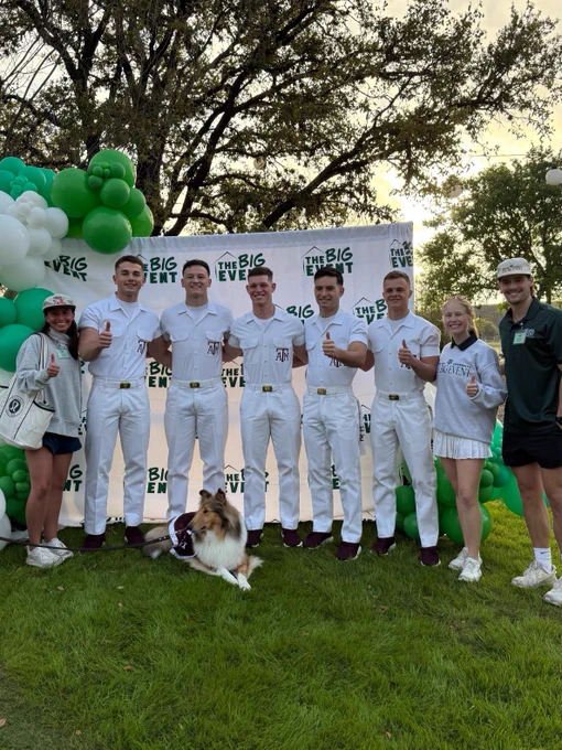 A group of people stands on grass in front of a backdrop with "The Big Event" logo. They are dressed in coordinated white and green outfits. Texas A&M University is associated with the event. Texas A&M mascot, Reveille, is sitting in front of them. Balloons are on the left side of the image. Trees are visible in the background.