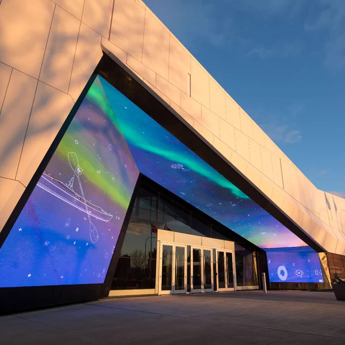 Entrée d'un bâtiment moderne avec de grands écrans numériques affichant des motifs abstraits colorés sous un ciel bleu clair.
