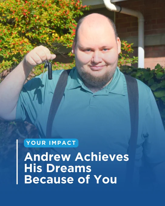 Andrew, a person supported by SJCS, stands outdoors smiling and holding up a set of car keys. He is wearing a teal polo shirt with suspenders, with a red berry bush and brick building visible in the background.
