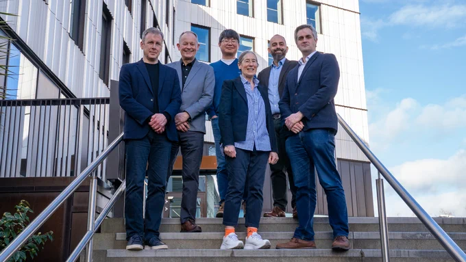 A group of six people, five men and one woman, stand on outdoor steps in front of a modern building. They are all dressed in business attire. 