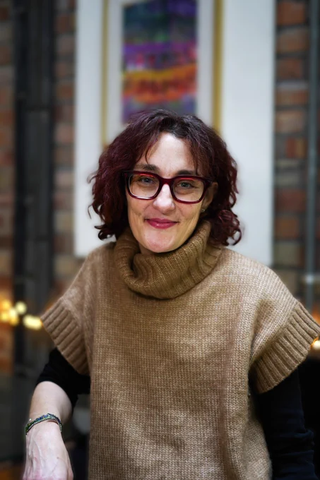 Caroline is a white woman with shoulder-length dark hair. She wears glasses and a polo neck wool knit with short sleeve. She sits inside a building. 
