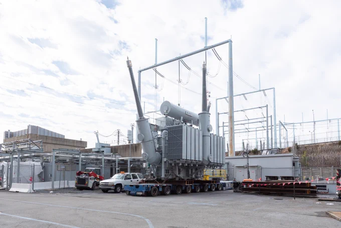 A large transformer is being transported on a flatbed trailer within a nuclear plant setting, surrounded by utility structures and power lines. Several vehicles are parked nearby under a partly cloudy sky.