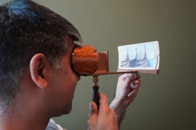 A man holds a brown viewfinder device by a handle, in front of his face. The viewfinder covers his eyes and is connected to a stereocard that the man holds with his other hand. The stereocard is held up vertically, so the man can view it.