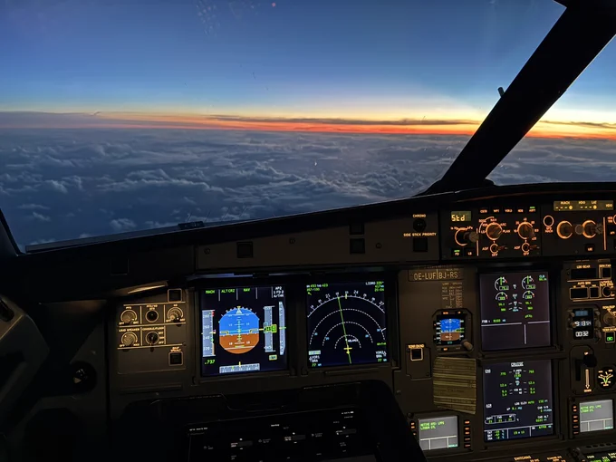 In cruise, view out of the flightdeck over clouds below and sunset colors on the horizon.