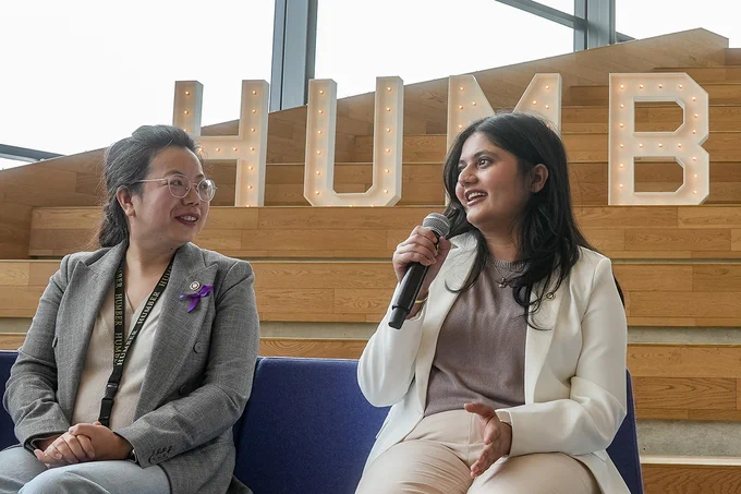 Two people sit together on a stage. One is speaking into a microphone.
