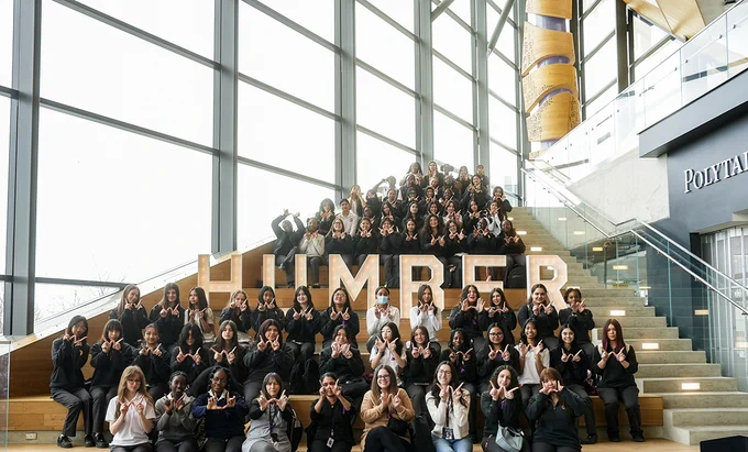 A group of people are sitting around a Humber sign while holding up their hands in the shape of a W.
