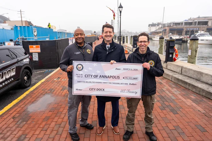 Annapolis OEM Director, Kevin Simmons, City of Annapolis Mayor, Jared Littman, and Annapolis OEM Deputy Director David Mandell stand along the City Dock waterfront holding an oversized check. The check is made out to the City of Annapolis for $33,522,069 from the U.S. Treasury. The memo reads "City Dock". Boats, buildings, and construction fencing are visible behind them.