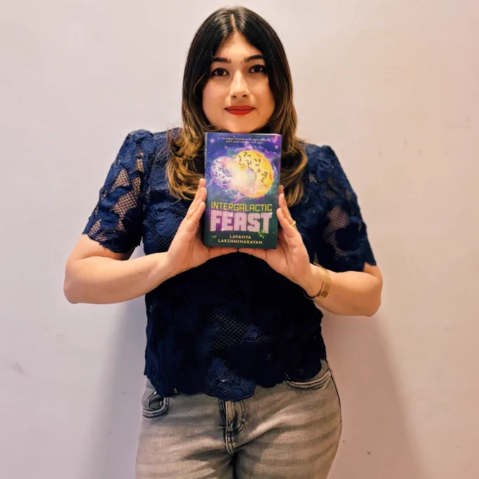 I’m standing against a plain light wall, holding a copy of Intergalactic Feast at chest height so the colorful space-themed cover faces the camera. I’ve got long, dark hair, and I’m wearing a navy lace top, light jeans, and red lipstick, looking directly at the camera with a small smile.