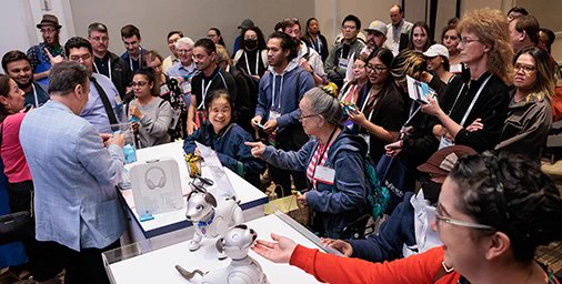 A large group of diverse people observe a demonstration at the C S U N Assistive Technology Conference Exhibit Hall.