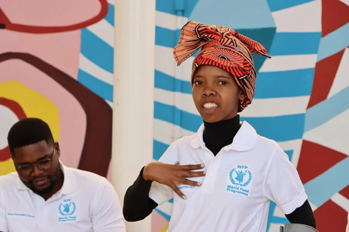 Delfin, a female WFP intern with a disability in Maputo, Mozambique, is shown speaking with one hand raised, wearing a white World Food Programme polo over a black turtleneck and a red-and-orange patterned headwrap; a male WFP staff member in a matching WFP polo sits to the side against a colorful painted mural backdrop. Photo credit: WFP/Ana Mato Hombre.