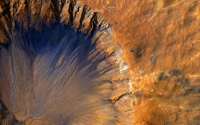 An impact crater with a sharp rim seen from above. Most of the landscape is orange, while the steep inner slope on the north side of the crater is a deep blue-black. Credit: NASA/JPL-Caltech/University of Arizona
