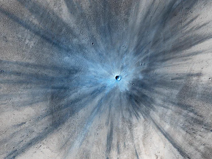 A fresh impact crater (at time of the image capture) is surrounded by a large blast zone with dark rays stretching outward from the crater. The crater itself looks light blue. Credit: NASA/JPL-Caltech/University of Arizona