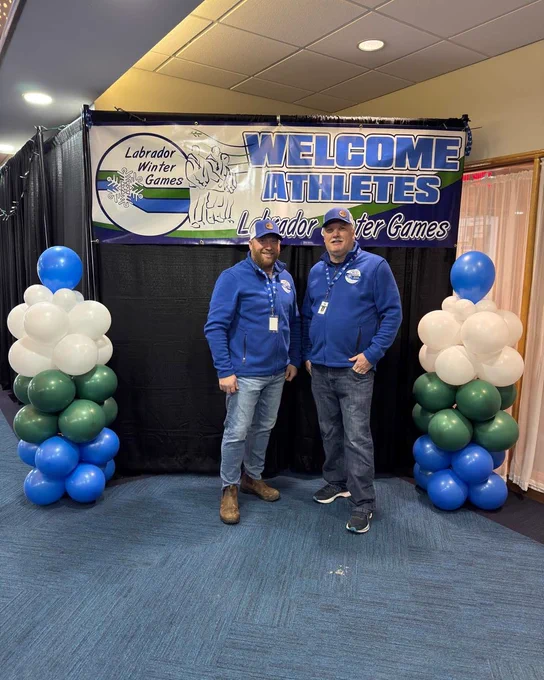Two men stand in front of a banner that reads “Welcome athletes”. They are wearing matching blue sweaters and baseball hats. 