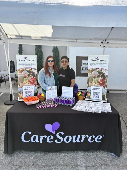 A promotional booth under a white canopy with a CareSource tablecloth. On the table are bowls of small basketballs, promotional items, and literature. Two people are standing behind the table, smiling. CareSource banners are displayed in the background.