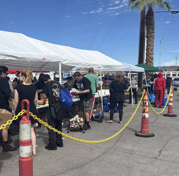 A community food distribution event is taking place outdoors under white tents. People are gathered around tables with various food items, such as cans and produce. Traffic cones and a yellow chain create a designated pathway. Palm trees and a partly cloudy sky are visible in the background.