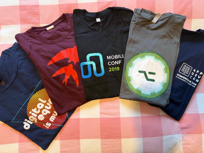 Five folded conference t-shirts laid out on a pink checkered surface. From left to right: a navy "Digital Equality" shirt, a burgundy shirt with the Swift birds, a black "Mobile Conf 2019" shirt, a grey shirt with the AltConf logo, and a navy "AppDevCon Conference 2018" shirt.