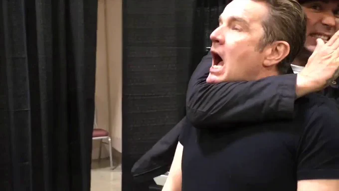 The image shows a man (James Marsters) in a black shirt with his mouth open wide, appearing to be in a playful or dramatic pose. Another person (Jason David Frank), wearing dark clothing and partially hidden, is standing behind him with an arm wrapped around the first man's throat. The setting appears to be an indoor event space with dark curtains and a glimpse of a red chair in the background.