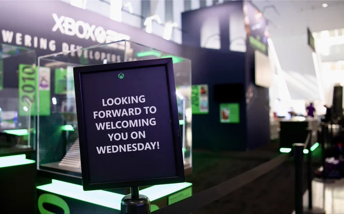 A black sign on a stand at the Xbox GDC exhibit reads, “Looking forward to welcoming you on Wednesday!” Green accent lighting surrounds the display, with Xbox signage in the background. (Alt generated by Microsoft Copilot and reviewed for accuracy)