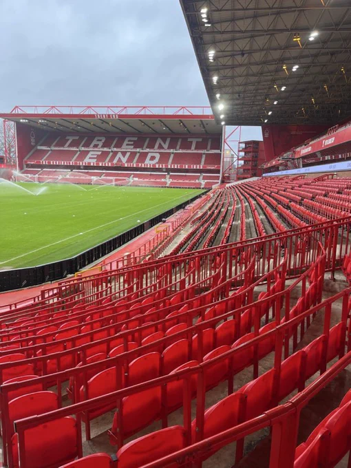 Photo of an empty City Ground, home of Nottingham Forest FC. 
The shot is taken at the very end of the Brian Clough stand lower tier, where it joins the Bridgford stand. The bottom third of image depicts curved rails and red seats which sweep round tho the right, joining the lower tier previously mentioned. Hovering above the lower tier a light grey roofing structure can be seen with a web of ironwork and some lighting in its underbelly. At the far top right of the photo you can just make out a stack of red shipping containers which are used as corporate suites, these are at the very opposite end of the Clough stand. 
Towards the top left of the image a bright green pitch is being watered by a series of automatic sprinklers in front of the double tier Trent End stand. The words “Trent End” are clearly visible in white seating in the top tier of this stand against the backdrop of the predominantly red seats. 
The photo was taken in daylight and shows a light blue/grey  sky above.