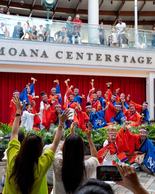 A group of performers in colorful traditional attire poses enthusiastically on a stage labeled "Moana Centerstage." They stand in front of a red curtain, holding fans and wearing patterned headbands. An audience is gathered, with some individuals taking photos. The stage area is decorated with greenery.
