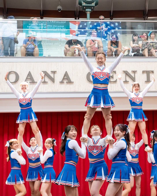 A cheerleading team performs a routine, forming a pyramid with several members lifted in the air. They are wearing blue outfits with "Sophia" written on the front. An audience observes from a higher level in the background at Ala Moana Center.