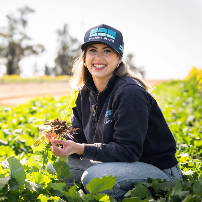 Sayra Samudio of Riverine Plains wearing a cap and dark jacket crouches in a green leafy field holding soil and plant roots. Post announcing her as an AgriFutures-sponsored participant in the 2026 ARLF TRAIL program.