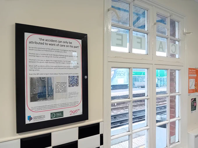Photograph of a poster in a frame, titled 'the accident can only be attributed to want of care on his part', detailing the life of goods porter Thomas Smith, on a waiting room wall, with a window beyond. Through the window the station platform and railway tracks can be seen.