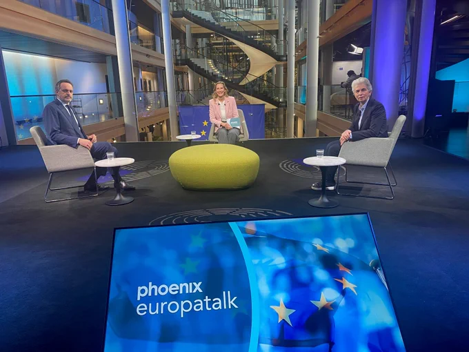 Eine Gruppe von drei Personen sitzt an einem Tisch in einem modernen Fernsehstudio mit dem Logo "Phoenix Europatalks".