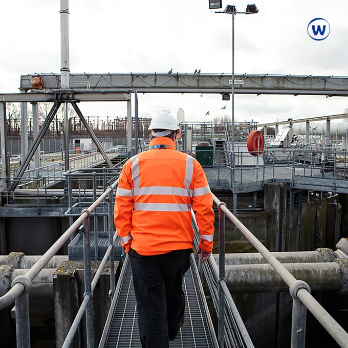 The photo is of a man wearing a white hard hat and an orange hi-vis jacket. He is outside. Canary Wharf is visible in the distance.
