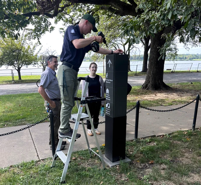 A man stands on a ladder using a drill to repair a sign next to a sidewalk in a park while a National Park Service worker and a young woman watch.
