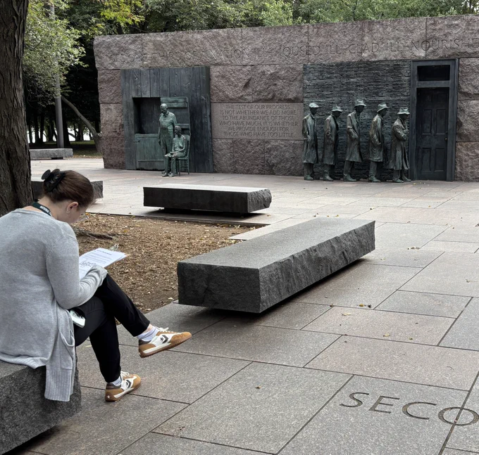 A young woman sits on a stone bench in an outdoor memorial drawing a sketch of two sculptures in front of her.