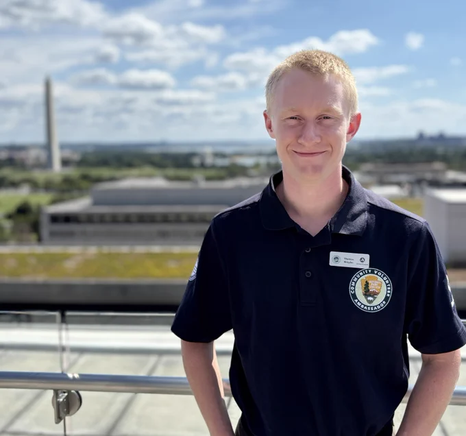 A smiling young man wearing a dark polo shirt with the National Park Service logo on the chest stands on a rooftop with the Washington Monument in the background.