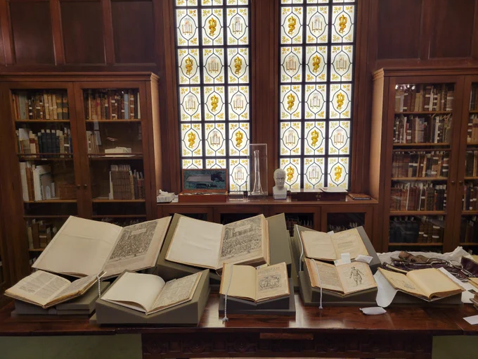 Image of a table in a wood-paneled library, with a tall painted glass window behind. There are 2 rows of books open on supports, three large works in the back, 4 smaller ones in the front. 