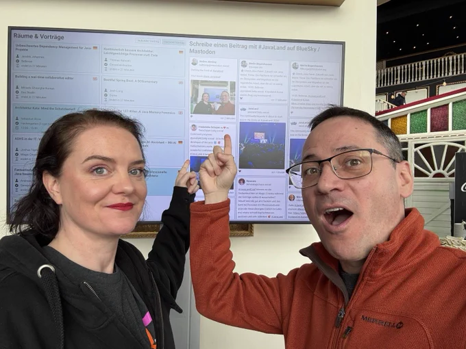 Marit van Dijk and Andres Almiray in front of a screen showing the JavaLand program and social media feed. They are both pointing at the feed, which shows a post with a picture of the both of them.