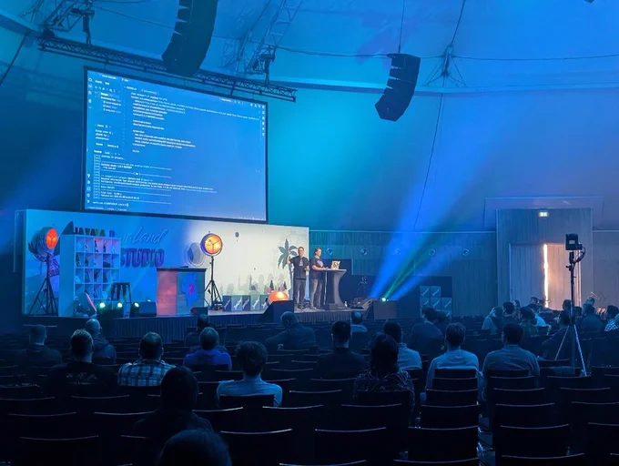 Andres Almiray and Marit van Dijk on stage at JavaLand. Andres is raising his hand, Marit is typing. The screen behind them shown Maven in IntelliJ IDEA. 