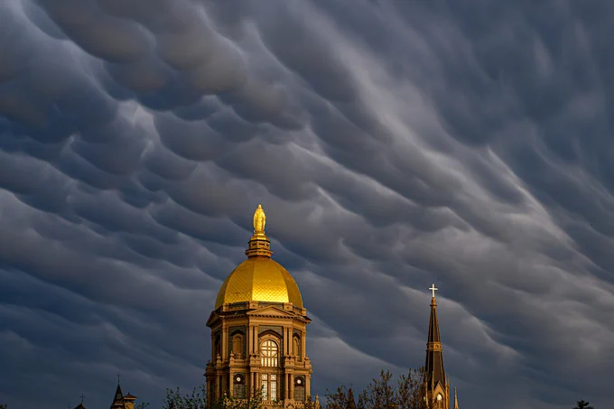 A dramatic sky with wavy clouds looms over the golden dome of the University of Notre Dame's Main Building, accompanied by a church spire with a cross.