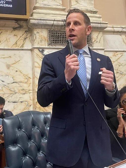 Delegate Edelson wearing a blue suit and tie holds a microphone and gives a speech 