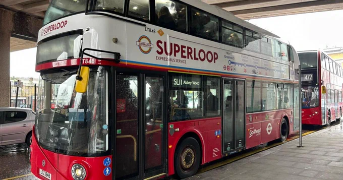 A red double‑decker bus branded for the Superloop express service is parked at a bus stand. The destination display reads “SL1 Abbey Wood,” and another red bus is visible behind it under the station canopy.